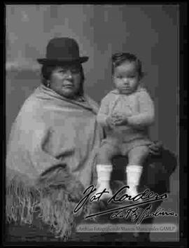 Foto de estudio de una madre parada junto a su niño pequeño, que se encuentra sentado en la cabecera de un sillón