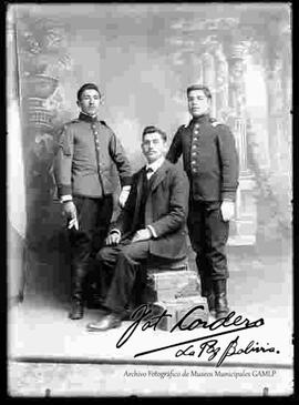 Foto de estudio de tres varones. Uno  vestido de traje y sentado en unos escalones en medio de dos jóvenes cadetes