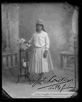 Foto de estudio de una chola que se encuentra parada y apoyada a una mesita con flores. Lleva puesto un sombrero borsalino de color oscuro, blusa de hilo, pollera y botines con tacón