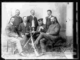 Foto de estudio de un grupo de jugadores de tiro al blanco, sentados en una mesa junto a fusiles y su trofeo