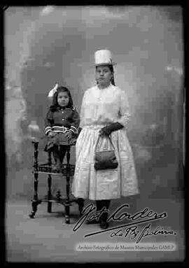 Foto de estudio de una chola que se encuentra parada y apoyada a una mesita donde esta una niña sentada. Lleva puesto un sombrero blanco de copa alta, una blusa de media manga con guantes, porta una cartera, pollera y botines con tacón.