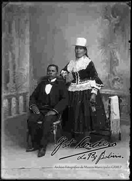 Foto de estudio de una pareja. Una chola que se encuentra parada junto a su esposo que esta sentado en una silla de mimbre. Lleva puesto un sombrero blanco de copa alta, una blusa de piel de lobo con encajes, pollera con figuras y botines con tacón. Sostiene en mano una pequeña escarcela (cartera).