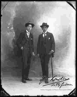 Foto de estudio de dos varones que visten de traje y sombrero. ambos portan en mano guantes y solo uno de ellos un paraguas.