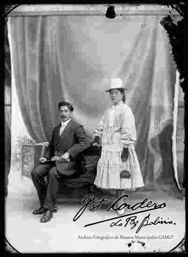 Foto de estudio de una pareja. Una chola junto a un varón de traje y corbata sentado en un sillón. Lleva puesto un sombrero blanco de copa alta, blusa suelta blanca de cuello cerrado, guantes,  pollera y  botines con tacón. Sostiene en mano una escarcela (cartera).