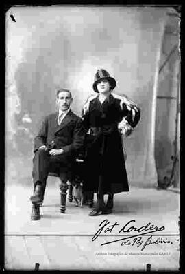 Foto de estudio de una pareja con su perrito. El varón se encuentra sentado en un sillón y lleva puesto un traje formal. La dama se encuentra parada y lleva puesto un sombrero de dos colores, un vestido y abrigo de gamuza con una estola de zorro.