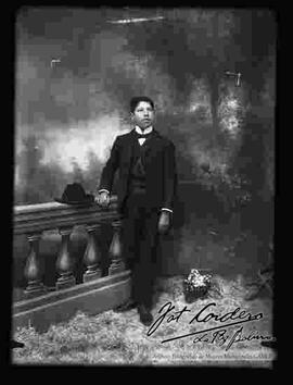Foto de estudio de un joven que se encuentra parado y apoyado a un barandal. Lleva puesto una camisa con cuello almidonado, corbata de gato y  un traje con chaleco de color negro.