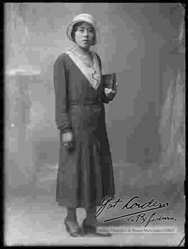 Foto de estudio de una dama. Lleva puesto un vestido largo y un sombrero, sostiene en mano una pequeña cartera.