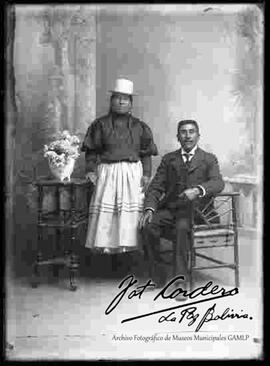 Foto de estudio de una pareja. Una chola que se encuentra parada y apoyada a una mesita con flores. Lleva puesto con sombrero de copa alta de color blanco, una blusa cerrada, pollera con enagua sobresalida y botines con tacón. A su lado se encuentra un caballero sentado en un banco vestido con traje y corbata