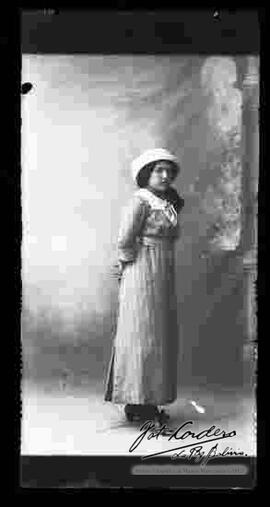 Foto de estudio de una joven señorita que lleva puesto: Un sombrero, una blusa, un vestido largo con botones y zapatos con tacón.