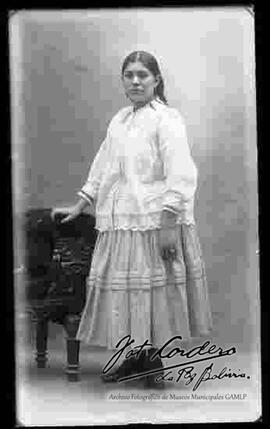 Foto de estudio de una chola sin sombrero y apoyada en un sillón. Lleva puesto una blusa blanca larga, pollera y botines con tacón