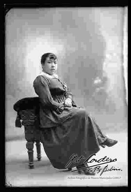 Foto de estudio de una señorita que se encuentra sentada en un sillón. Lleva puesto un vestido o traje formal y botines con tacón.