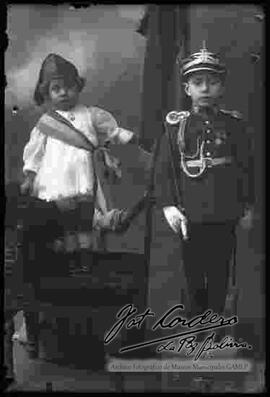 Foto de estudio de dos niños. Un niño vestido de militar y niña con una banda, representando a la madre patria