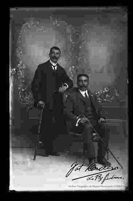 Foto de estudio de dos varones de bigote que visten de traje.