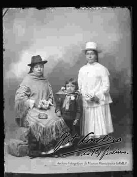 Foto de estudio de dos cholas junto a una niña. Una de ellas se encuentra parada y viste de blanco, lleva puesto un sombrero blanco de copa alta, blusa suelta de mangas largas y con encaje, pollera y botines con tacón. La otra se encuentra sentada y lleva puesto un sombrero de color oscuro, una manta bordada, pollera y botines con tacón, sostiene en mano una escarcela (cartera) y flores