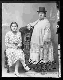 Foto de estudio de una chola que se encuentra parada junto a su hija que que esta sentada en un sillón. Lleva puesto un sombrero borsalino de color oscuro, manta bordada, pollera a rayas y botines con tacón