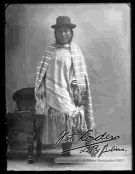 Foto de estudio de una chola de provincia apoyada a un sillon. Luce un sombrero borsalino de color negro, manta a rayas, pollera y zapatillas planas
