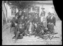 Grupo de varones (amigos) vestidos de traje y sombrero. Se encuentran reunidos en el patio de una casa particular