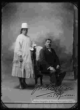 Foto de estudio de una pareja de esposos. Una chola que se encuentra parada, con sombrero blanco de copa alta, manta, pollera y botines con tacón. Su esposo viste de traje y corbata que esta sentado en un sillón donde esta puesto una manta bordada