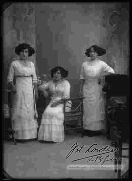 Foto de estudio de tres damas (hermanas). Llevan puesto vestidos de fiesta largos, de color claro y adornados con encajes.