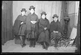 Foto de estudio de cuatro cholas. Tres se encuentran sentadas y una parada. Llevan puesto sombreros borsalinos negros, mantas tejidas de color oscuro, polleras y zapatillas