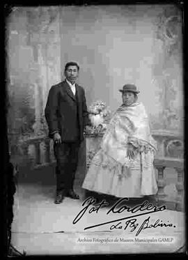 Foto de estudio de una pareja de provincia. Una chola apoyada a la columna de una baranda con flores. Lleva puesto un sombrero borsalino, manta tejida, pollera, zapatillas. Se encuentra sentada  junto a un varón  de traje oscuro