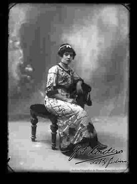 Foto de estudio de una señorita que se encuentra sentada en un sillón. Lleva puesto una wincha de perlas en el cabello,  un vestido de fiesta adornado y botines con tacón. Presenta asi mismo unos aretes, una gargantilla, un collar de perlas y anillos en la mano.