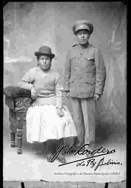 Foto de estudio de una pareja de esposos. Una chola que se encuentra sentada en un sillón, luciendo un sombrero borsalino de color oscuro, chompa cerrada, pollera y zapatillas planas. Su esposo se encuentra parado y vestido de policía