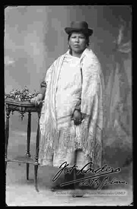 Foto de estudio de una chola que se encuentra parada y apoyada a una mesita. Luce un sombrero borsalino color negro, manta bordada prendida con un topo (prendedor), pollera y zapatillas planas