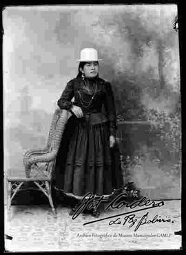 Foto de estudio de una chola que se encuentra parada y apoyada a una silla de mimbre. Lleva puesto un sombrero blanco de copa alta, una blusa negra cerrada de manga larga, joyas, pollera de color oscuro con centros sobresalidos y botines con tacón.