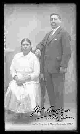 Foto de estudio de una pareja. Una chola que se encuentra sentada en un sillón a lado de su esposo y sin sombrero. Lleva puesto una blusa de media manga, falda larga y zapatillas con tacón