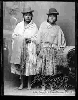 Foto estudio de dos cholas  paradas. Una sostiene en  brazo una manta y la otra se encuentra apoyada en un sillón donde esta puesto una manta. Ambas llevan puesto sombreros borsalinos de color oscuro, manta y botines con tacón.