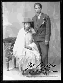 Foto de estudio de una pareja. Una chola que se encuentra sentada en un sillón, luciendo un sombrero borsalino de color negro, manta puesta, pollera y zapatillas planas. El caballero se encuentra parado y viste de traje y corbata