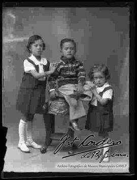 Foto de estudio de tres niños. Dos niñas y un niño que se encuentra sentado en un taburete