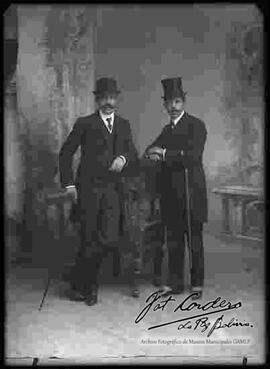 Foto de estudio de dos varones de bigote que visten de traje y sombrero.
