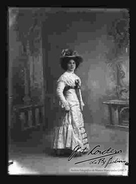 Foto de estudio de una dama que lleva puesto: Un sombrero adornado con flores, un vestido de fiesta con encajes, y guantes en las manos.