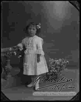 Foto de estudio de una niña que sostiene en mano unas flores y que se encuentra parada junto a un barandal