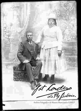 Foto de estudio de una pareja. Un caballero de traje y corbata sentado en un sillón junto a su esposa, una chola que se encuentra parada junto a él. Lleva puesto un sombrero blanco de copa alta, una chompa tejida de lana, pollera y botines con tacón.