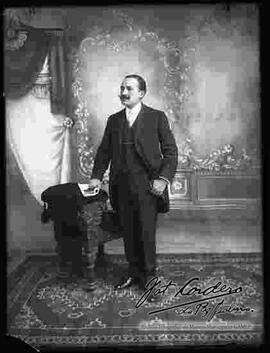 Foto de estudio de un varón que se encuentra parado a lado de un sillón. Lleva puesto una camisa blanca, corbata, chaleco y traje frac. Sostiene en mano unos guantes y una revista