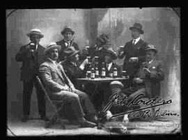 Foto de estudio de un grupo de varones vestidos de traje y sombrero, compartiendo unas cervezas