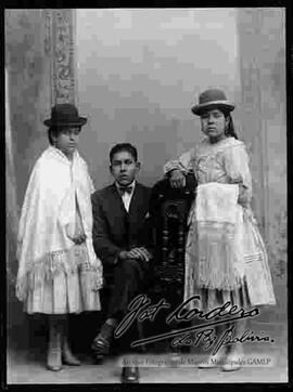 Foto de estudio de dos cholas que lucen sombreros borsalinos. Una se encuentra parada con manta puesta, la otra con manta en mano y pollera. Ambas llevan botines de tacón y estan junto a un caballero de traje que se encuentra sentado en medio de las dos