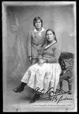 Foto de estudio de  una chola sin sombrero y sentada junto a un joven. Lleva puesto una chompa, pollera floreada y botines con tacón.
