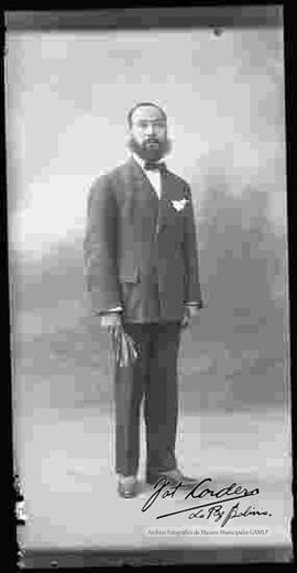 Foto de estudio de un varón de barba y bigote que lleva puesto: Un traje formal y sostiene en mano unos guantes.