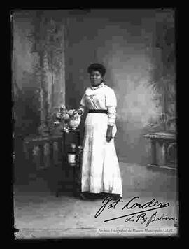 Foto de estudio de una dama que se encuentra parada junto a una mesita con un jarrón de flores. Lleva puesto un vestido largo con encajes, de color blanco y  sujeto por la cintura con un cinturón de color oscuro.