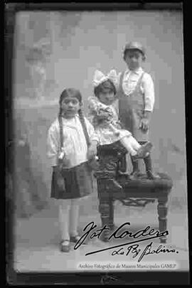 Foto de estudio de tres niños (hermanitos). Dos niñas y un niño parado encima de un sillón