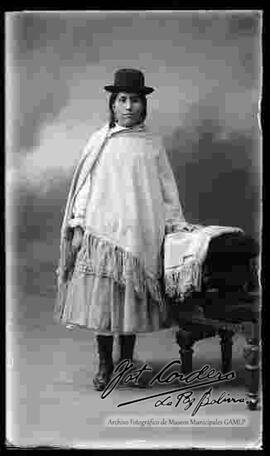 Foto de estudio de una chola que se encuentra parada y apoyada en un sillon donde esta su manta. Lleva puesto un sombrero borsalino de color negro, manta, pollera y botines con tacon