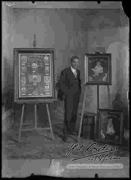 Foto de estudio de un pintor a lado de tres cuadros. Símbolos y emblemas de la patria y dos angelitos.