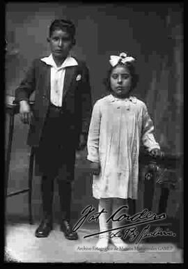 Foto de estudio de n niño y una niña (hermanos), parados a lado de muebles.