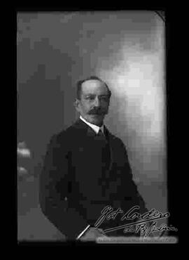 Foto de estudio de un varón de bigote que viste de traje y que se encuentra sentado.