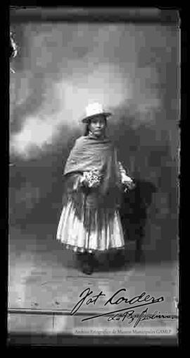 Foto de estudio de una chola que se encuentra parada y apoyada en un sillón. Lleva puesto un  sombrero blanco de copa alta, manta, pollera y botines con tacón. Sostiene en mano unas  flores