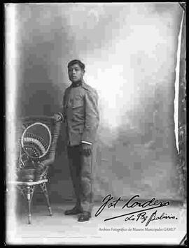 Foto de estudio de un soldado, con la mano apoyada a una silla de mimbre